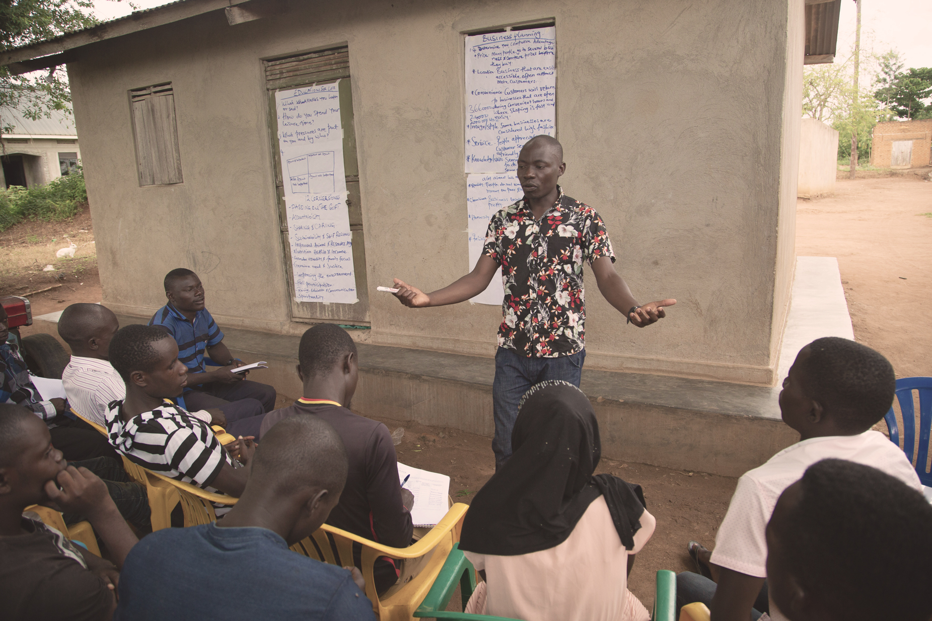 A young man in a Hawaiian shirt addresses members of the East Africa Youth Inclusion Project, who sit in plastic chairs in front of a small building with big pieces of paper taped to it that has notes about business development. 