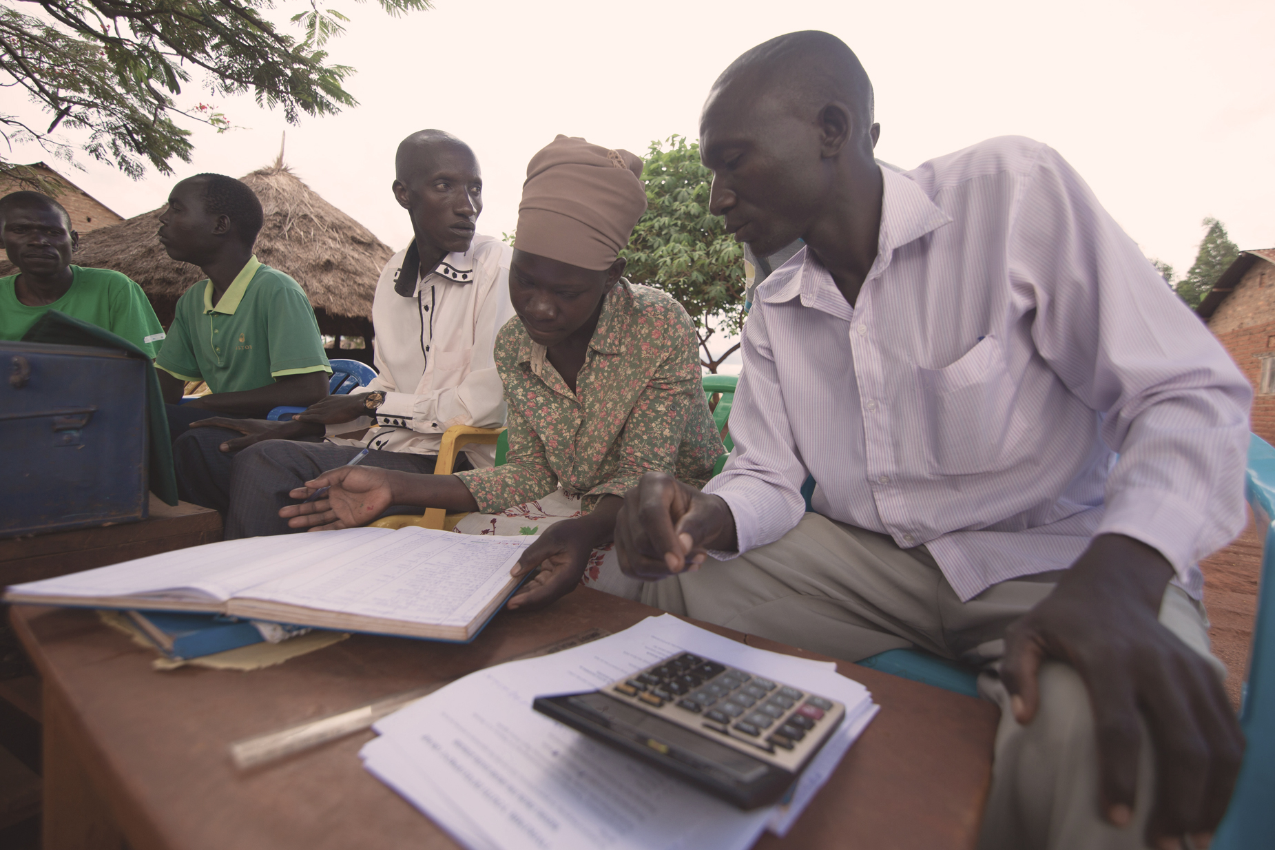 A young woman and a young man in the East Africa Youth Inclusion Project sit outside as part of a group. The two of them are looking over documents and have a calculator close at hand. 
