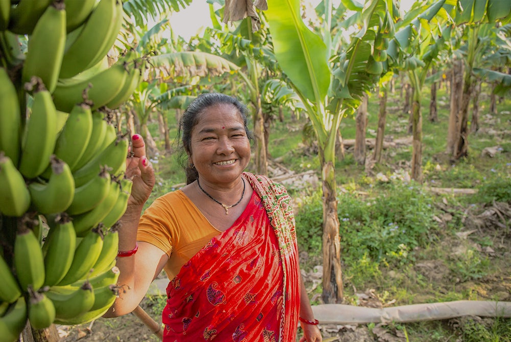 A woman in an orange sari smiles next to a banana plant.