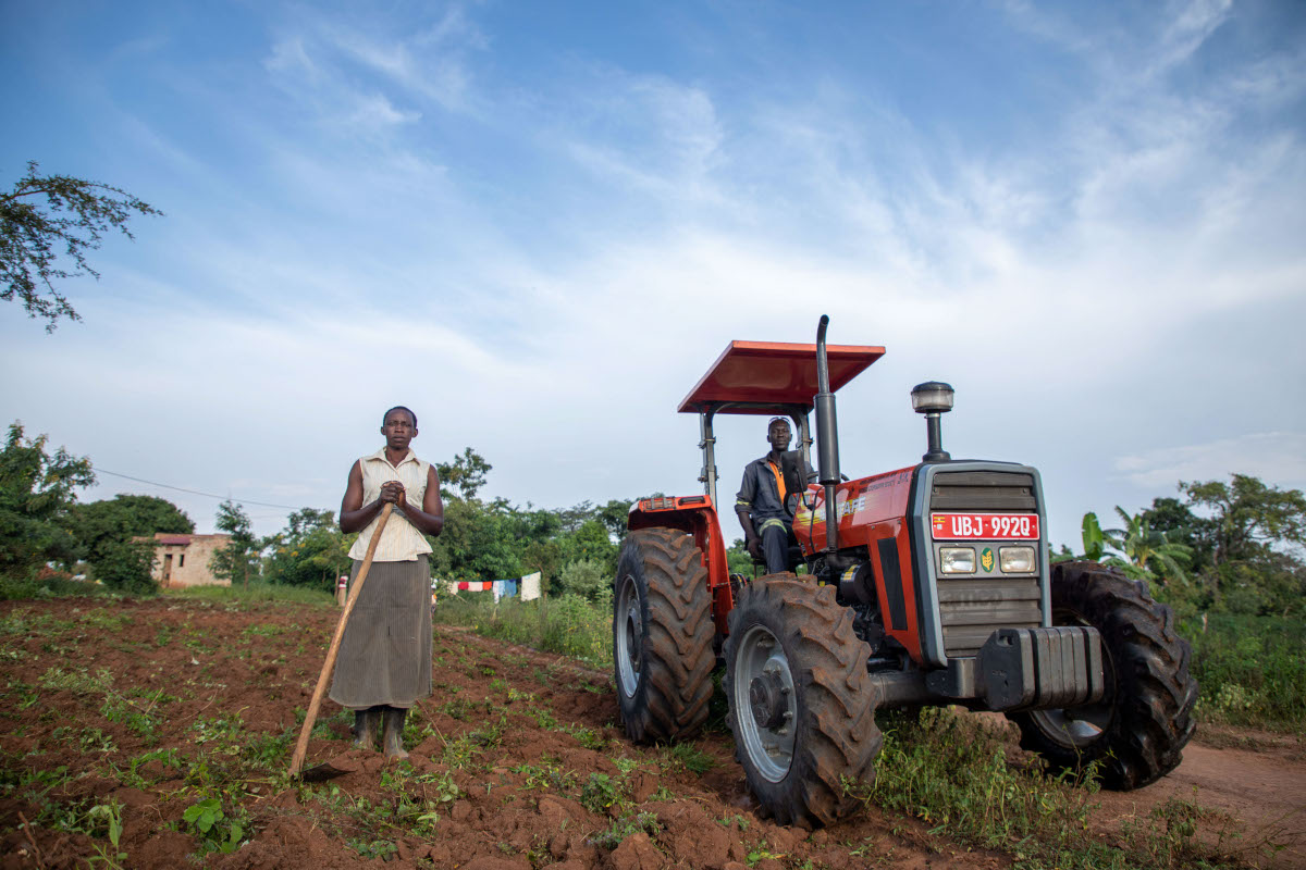 A woman holding a hoe and standing beside a tractor being driven by her husband in their field.