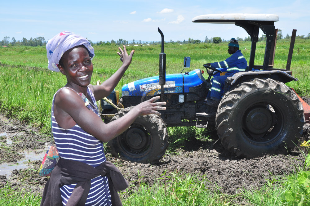 A woman stands in front of a tractor working in the background.