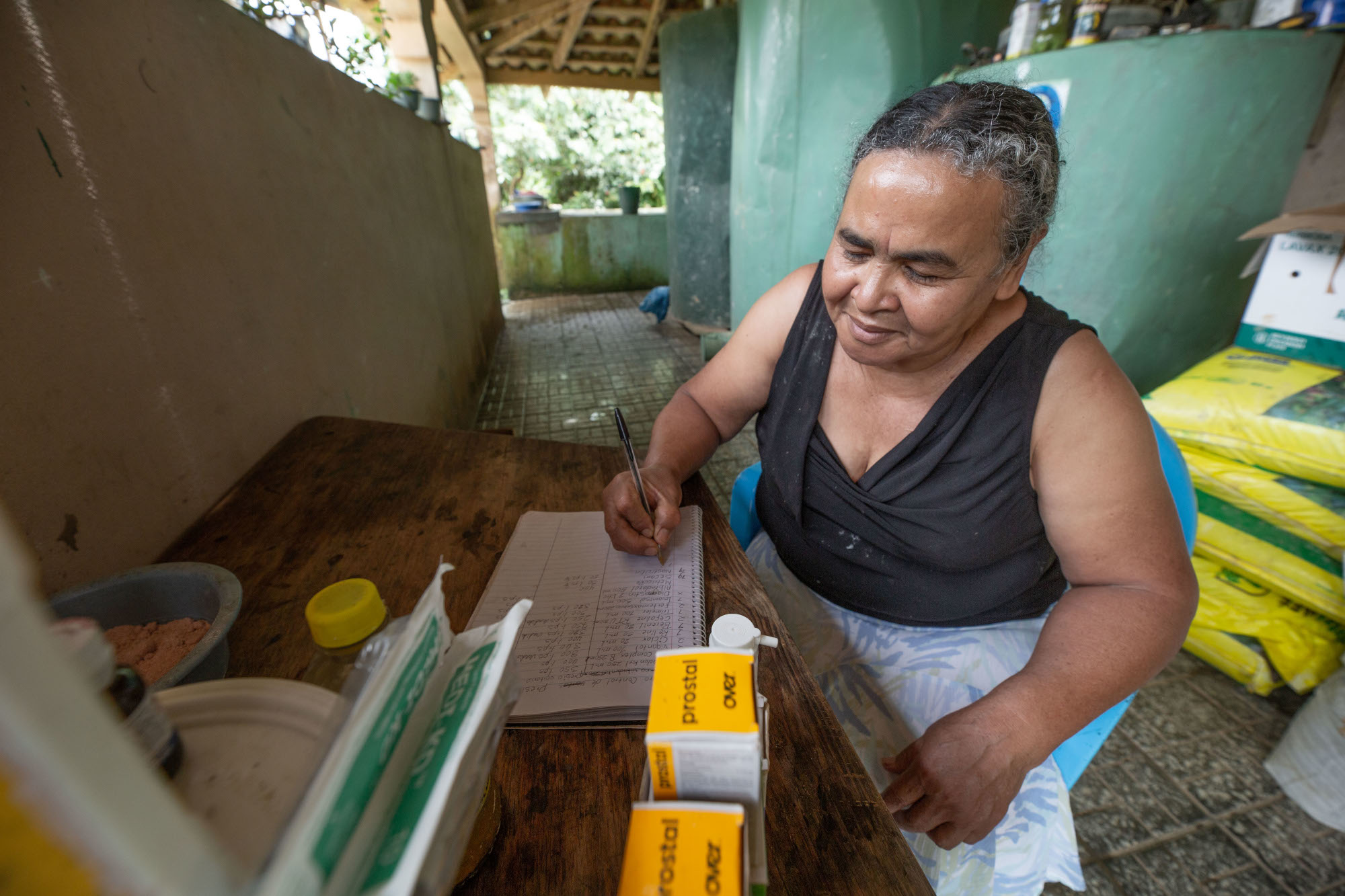 A woman entering her business records in a diary while sitting in a store where agricultural inputs are kept. 