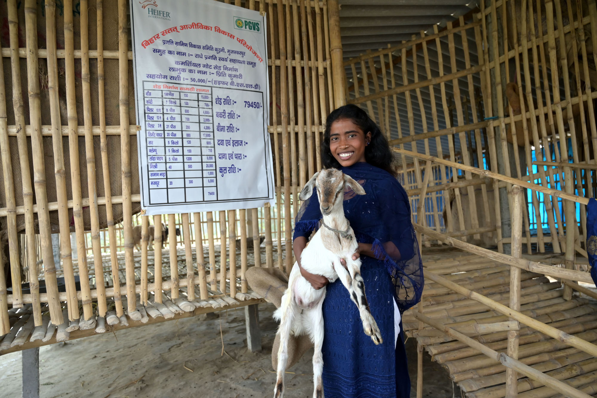 A woman holding a goat and standing in front of a newly constructed goat shelter.