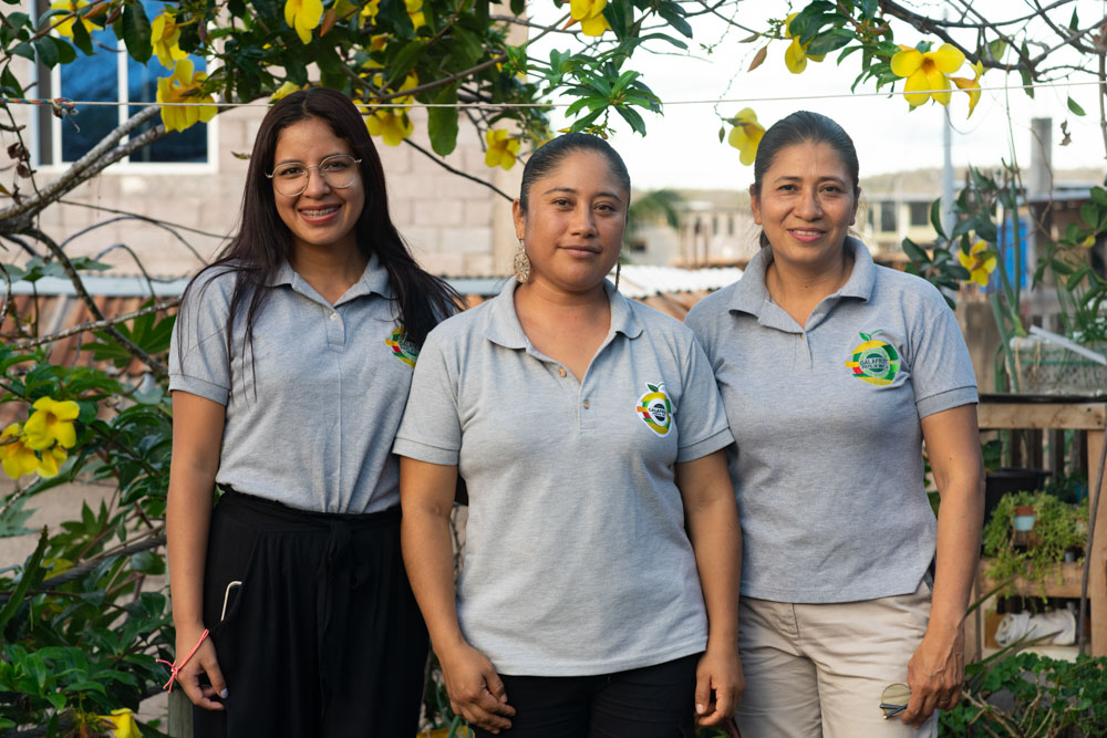 Three smiling women in gray polo shirts stand in front of yellow flowers.