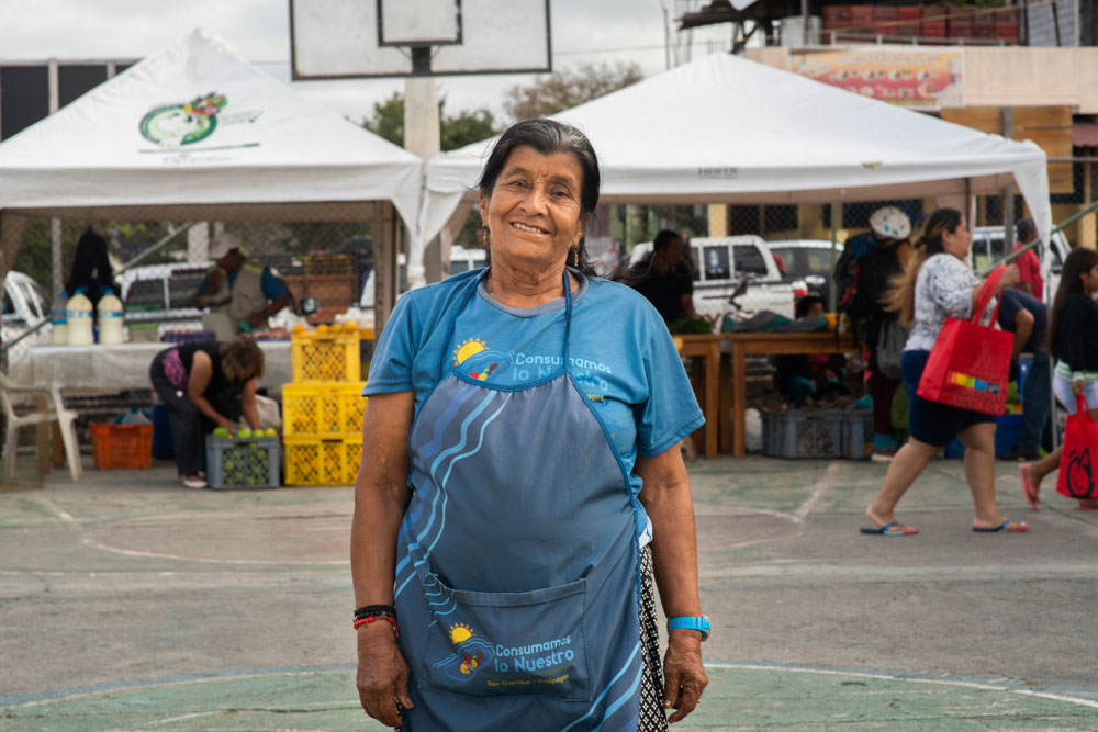 A smiling woman in a blue apron stands in a bustling outdoor market.