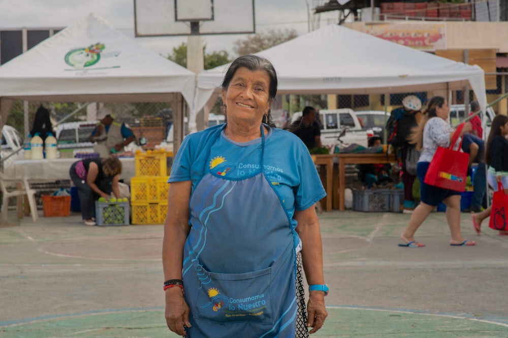A smiling woman in a blue apron stands in a bustling outdoor market.