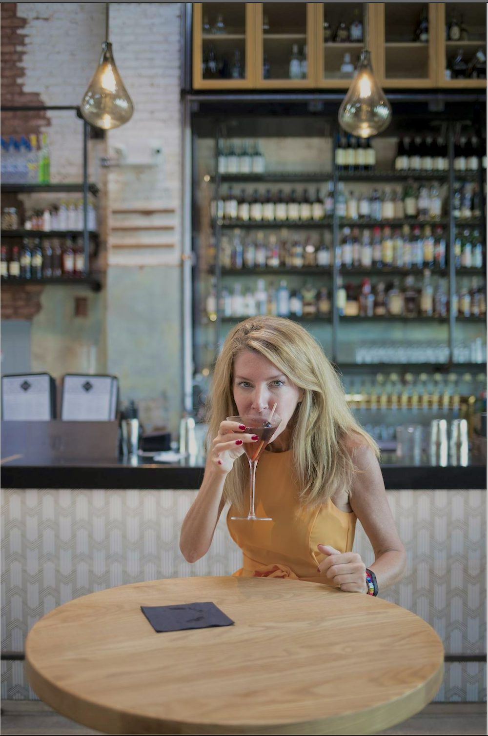 Laura Reiley sits at a bar table sipping a cocktail. She is wearing a yellow dress and looking directly into the camera over the rim of her glass. 